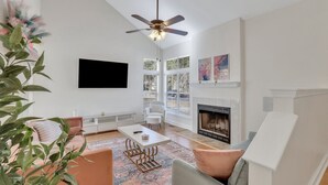 Una sala de estar moderna con chimenea, ventilador de techo, televisor de pantalla plana y ventanas amplias.