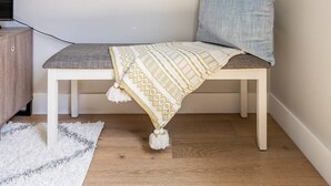 Una habitación acogedora con pared blanca, suelo de madera, un banco con un plaid estampado, almohada azul y un adorno blanco en la pared.