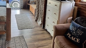 Interior acogedor de una cabaña con techo de madera, un televisor sobre un aparador, un sofá con un cojín y una alfombra estampada.