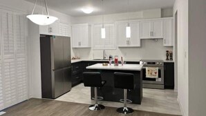 Moderni keittiö, jossa on valkoiset kaapit, keskellä oleva saareke baarituoleineen, ruostumattomasta teräksestä valmistetut kodinkoneet ja riippuvalaisimet.