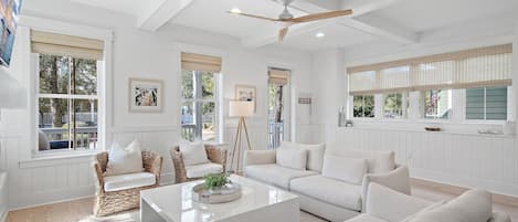 A spacious living room with white furniture, large windows, and a ceiling fan.