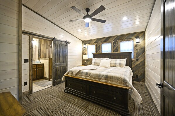 A bedroom with a wooden ceiling, a bed with a plaid bedspread, a nightstand, and a wall-mounted lamp.