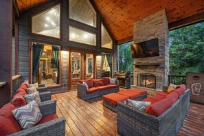 A wooden deck with wicker furniture, a stone fireplace, and large windows.
