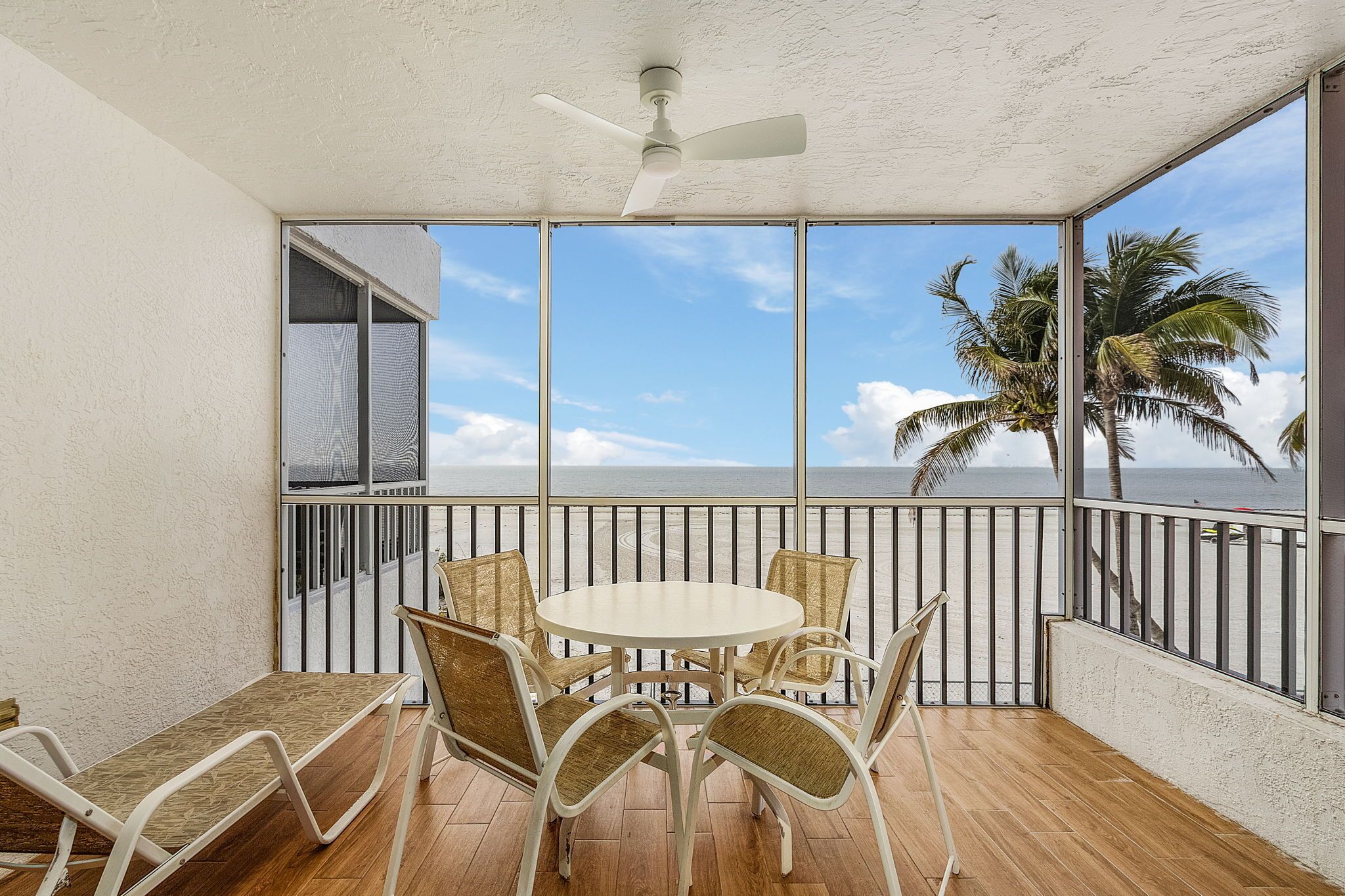 A balcony with a table and chairs, a ceiling fan, and a view of the ocean and palm trees.