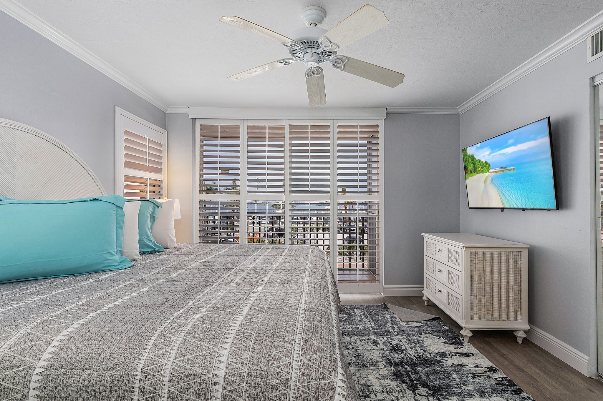 A bedroom with a bed, a ceiling fan, a TV, a dresser, and a view of a beach through the window.