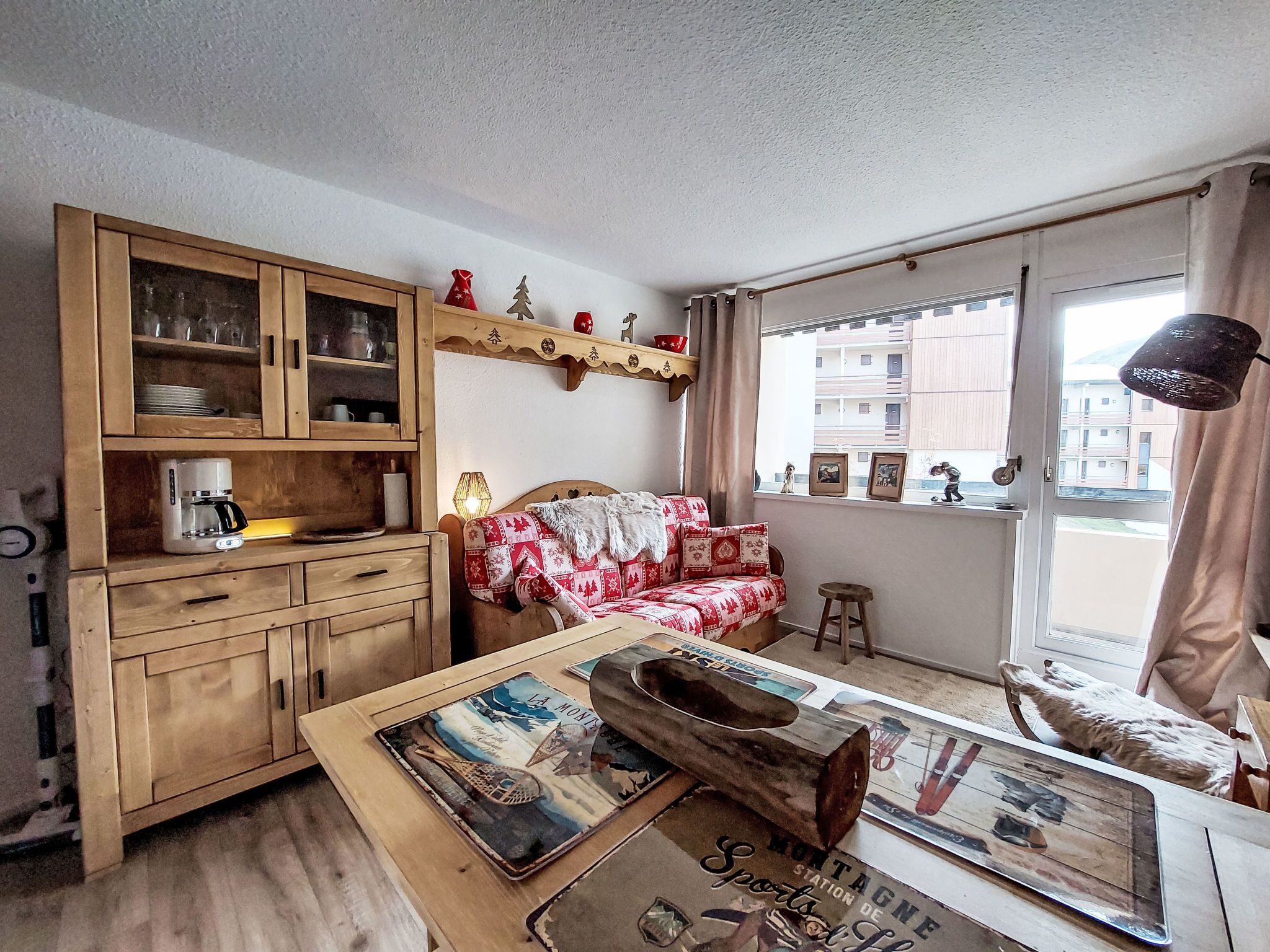 Ein gemütliches Zimmer mit einem Holzkasten, einem Sofa mit roter und weißer Decke, einer Kaffeemaschine und einem Fenster mit Blick auf Gebäude.