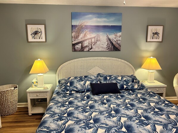 A bedroom with a bed, two bedside tables with lamps, a wicker chair, a woven basket, and a painting of a beach scene on the wall.