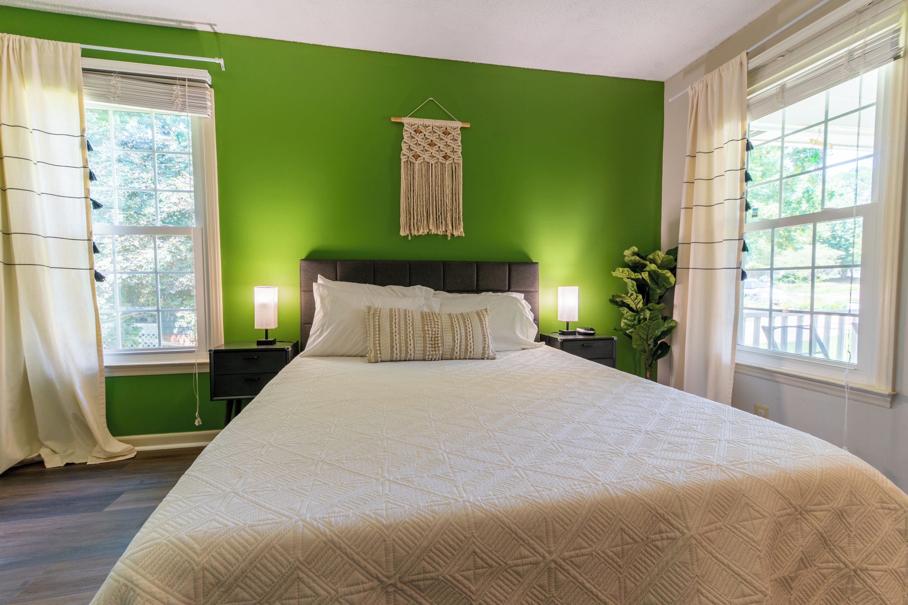 Een netjes opgemaakt bed met een geborduurd sprei, twee nachtkastjes met lampen, een macramé wanddecoratie, en uitzicht op groen door de ramen.