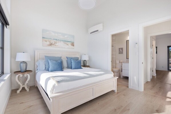 A bedroom with a large bed, two bedside tables, a nightstand, a painting on the wall, and an air conditioning unit.
