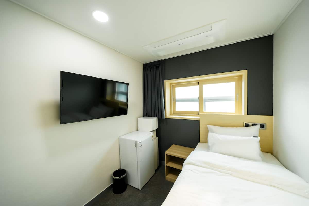 A hotel room with a bed, a TV mounted on the wall, a small desk, a window, and a wall-mounted air conditioning unit.