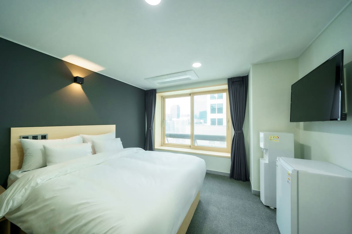 A hotel room with a bed, a flat-screen TV, a mini-fridge, and a window with curtains.