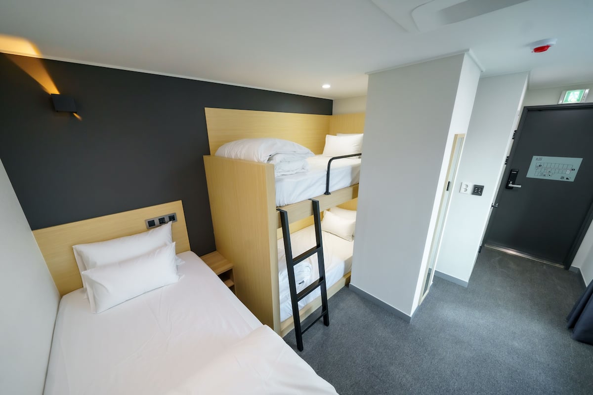 A hotel room with a bunk bed, a single bed, a desk, and a door leading to another room.