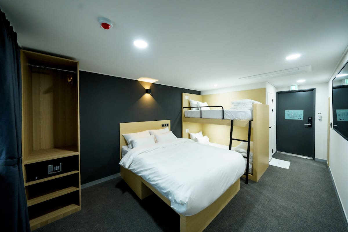 A hotel room with a bunk bed, a wardrobe with a control panel, a bedside table, and a door with a control panel.