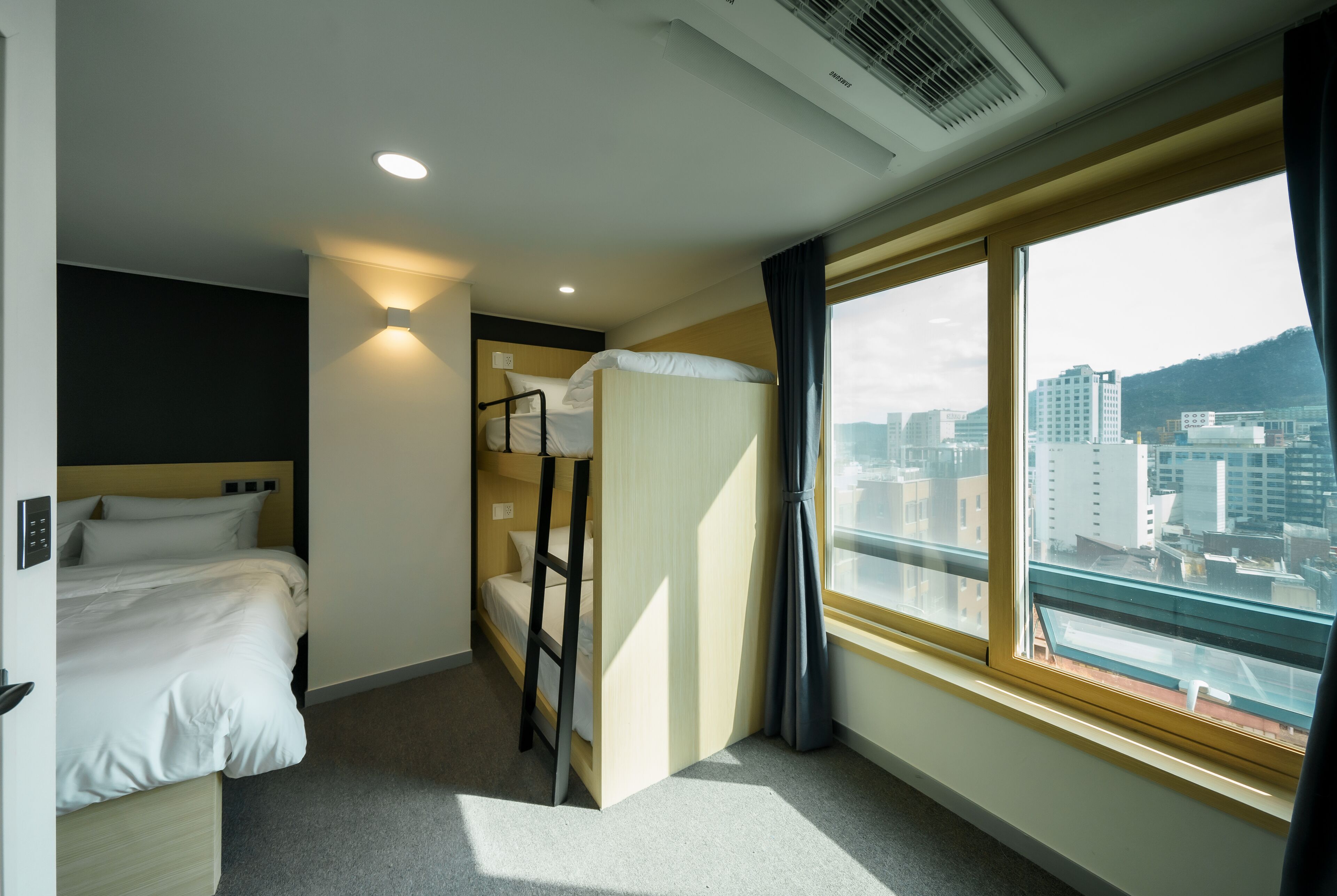 Habitación de hotel con literas, una cama individual, ventana con vista a la ciudad y un respiradero en el techo.