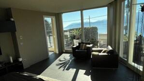 Une chambre d’hôtel moderne, dotée d’un grand lit, d’un canapé et d’un fauteuil. Une porte-fenêtre coulissante donne accès à un balcon offrant une vue sur les montagnes et un plan d’eau.