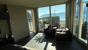 Een moderne hotelkamer met een groot bed, een bank en een stoel. Er is een schuifdeur naar een balkon met uitzicht op bergen en een wateroppervlak.