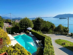 A swimming pool with lounge chairs, surrounded by greenery, with a view of a lake and mountains in the distance.