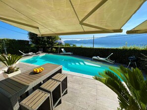 Ett poolområde med solstolar och ett bord, omgivet av en häck och med utsikt över havet.