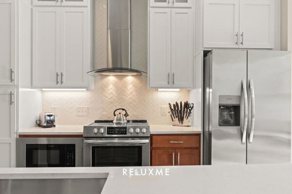 A modern kitchen with white cabinetry, a stainless steel range hood, a built-in oven, a microwave, and a refrigerator.