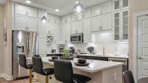 A modern kitchen with white cabinetry, stainless steel appliances, and black bar stools. - Reluxme | 2BR Condo in Center City w/ Spa Access (Charlotte)