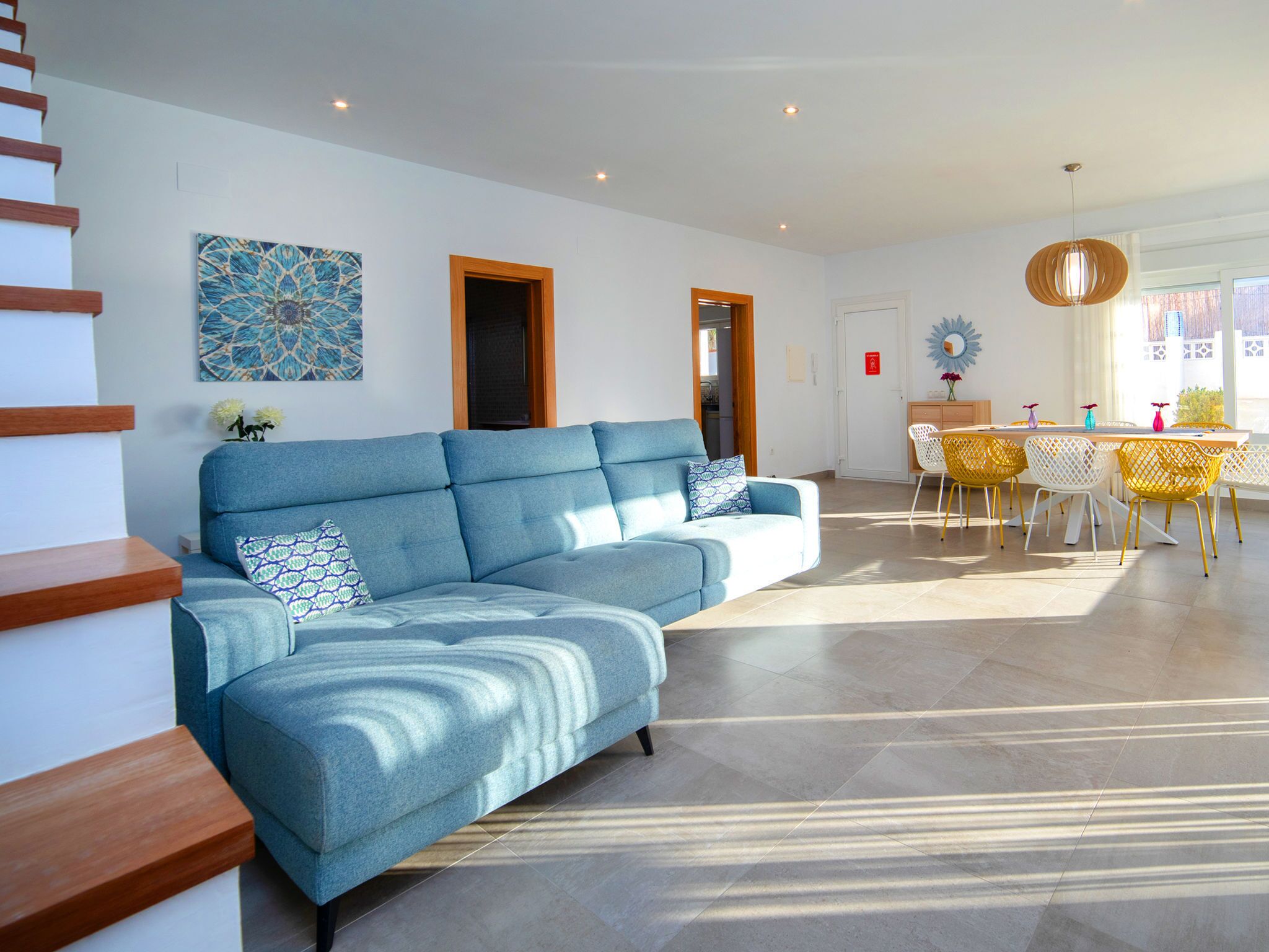 Ein modernes Wohnzimmer mit einem blauen Ecksofa, einem Essbereich mit gelben Stühlen und einer Treppe mit Holzstufen.