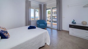 Ein Hotelzimmer mit Bett, Sessel, Kommode und Blick auf Strand und Gebäude.