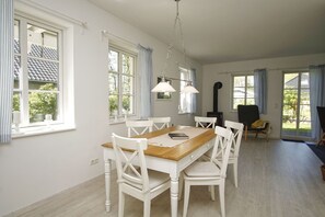 Ein Essbereich mit Holztisch und weißen Stühlen, großen Fenstern mit Vorhängen sowie einer hängenden Leuchte.