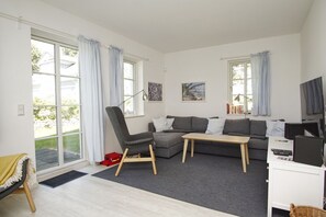 Uma sala de estar moderna com um sofá seccional cinza, uma mesa de centro de madeira e uma poltrona cinza. Há duas janelas com cortinas, uma televisão e uma estante.