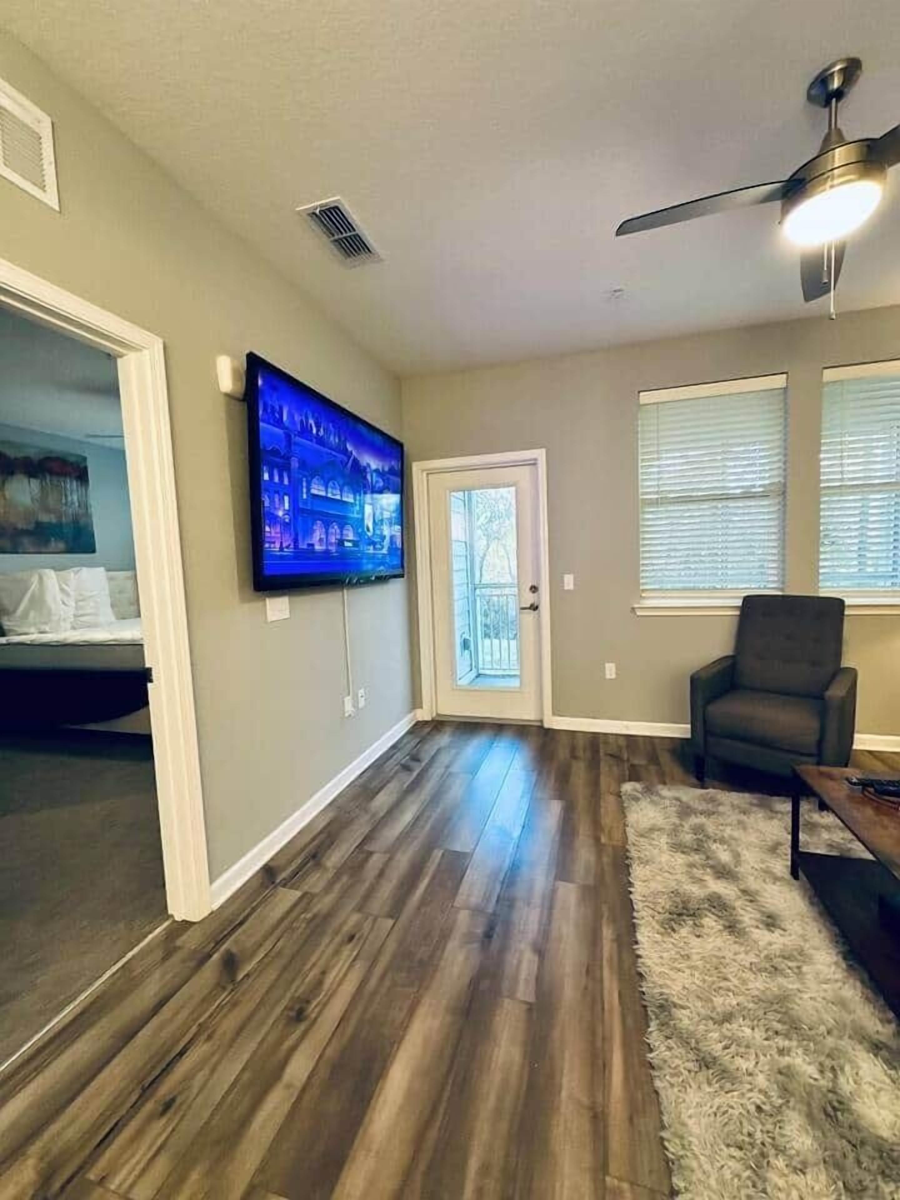 Una sala de estar con piso de madera, ventilador de techo, televisor de pantalla plana y puerta que da a un balcón.