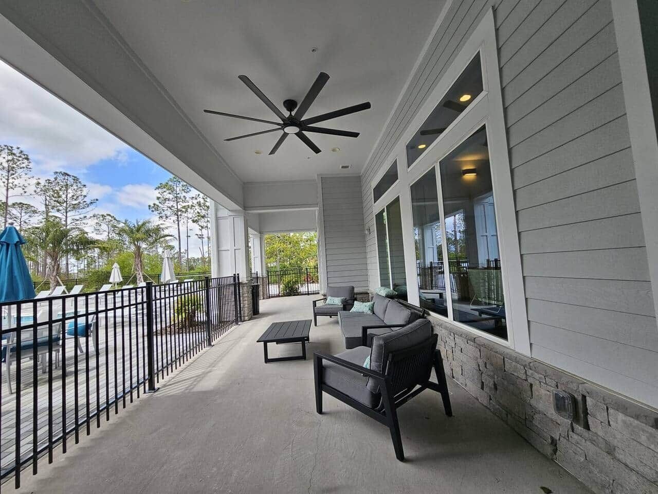 A covered outdoor seating area with a ceiling fan, a stone wall, and a view of the pool and trees.