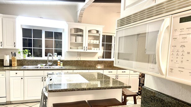 Una cocina moderna con gabinetes blancos, encimeras de granito, microondas integrado y un amplio ventanal.