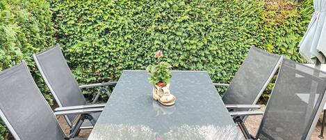 Un patio con una mesa y sillas, una planta en maceta y un seto al fondo.