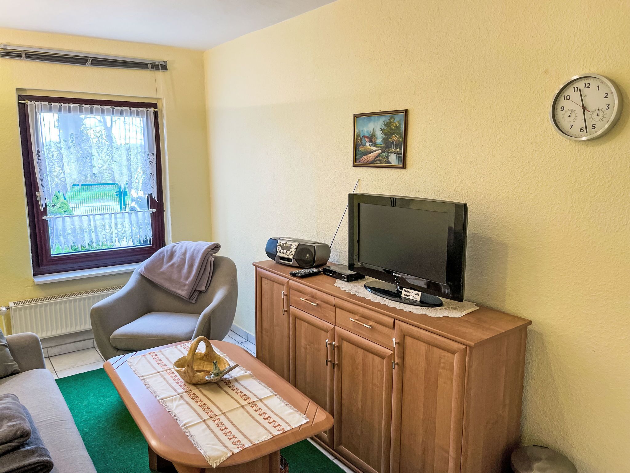 A room with a television on a wooden cabinet, a chair, a table with a placemat, a clock on the wall, and a window with a view of greenery.