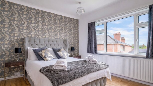 Une chambre à coucher avec une tête de lit capitonnée, une grande fenêtre avec des rideaux, un plancher en bois et un papier peint à motifs.