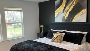 A bedroom with a large bed, a headboard, a nightstand, a lamp, and a window.