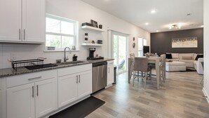 A modern kitchen with white cabinets, a dishwasher, and a coffee maker. There is a dining area with a table and chairs, and a living room with a couch and a painting on the wall.