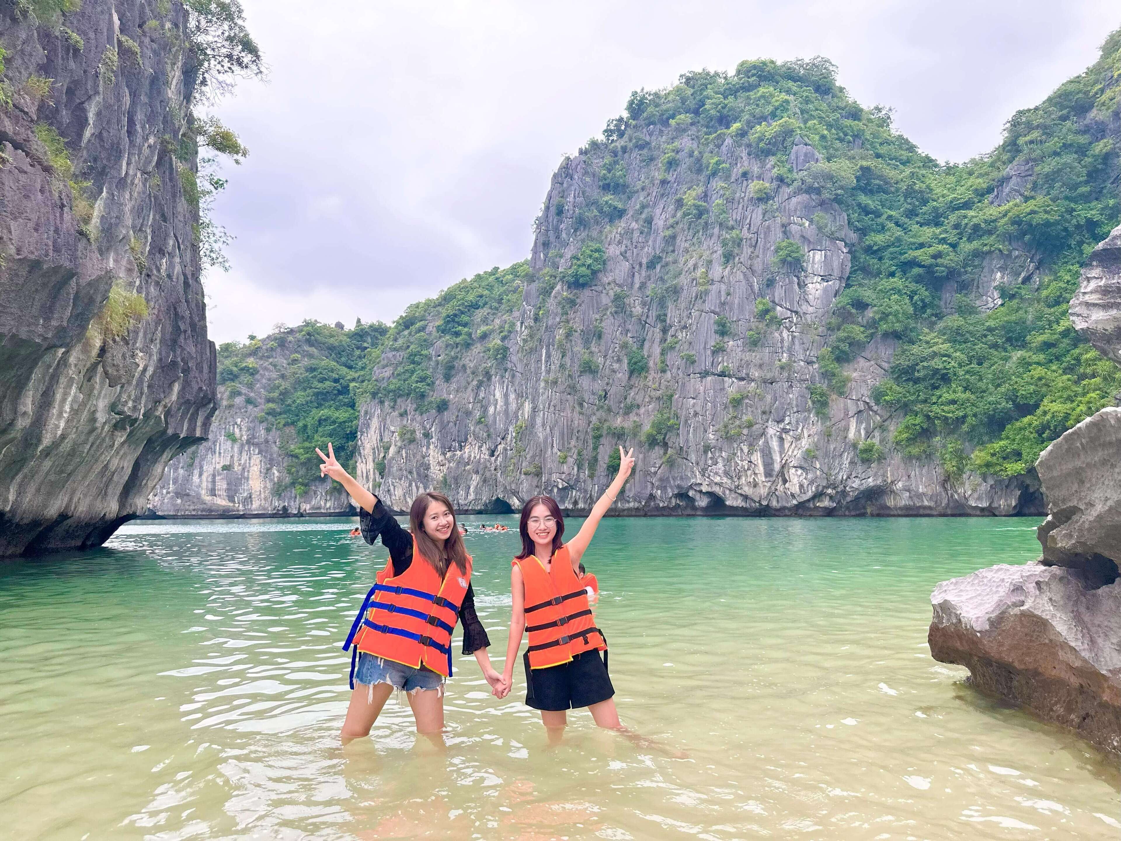 Two people in a clear water body with rocky cliffs in the background.