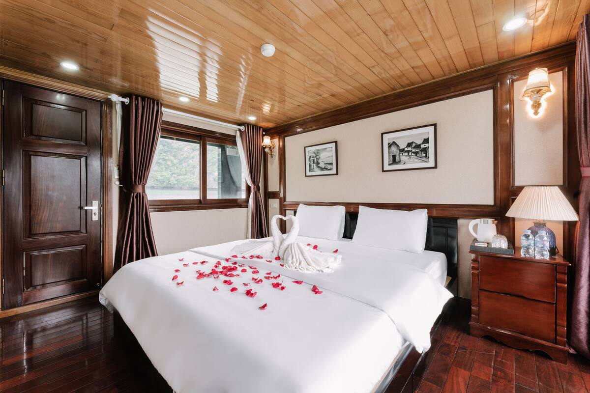A hotel room with a wooden ceiling, a large bed with white linens, a nightstand with a lamp, and two framed pictures on the wall.