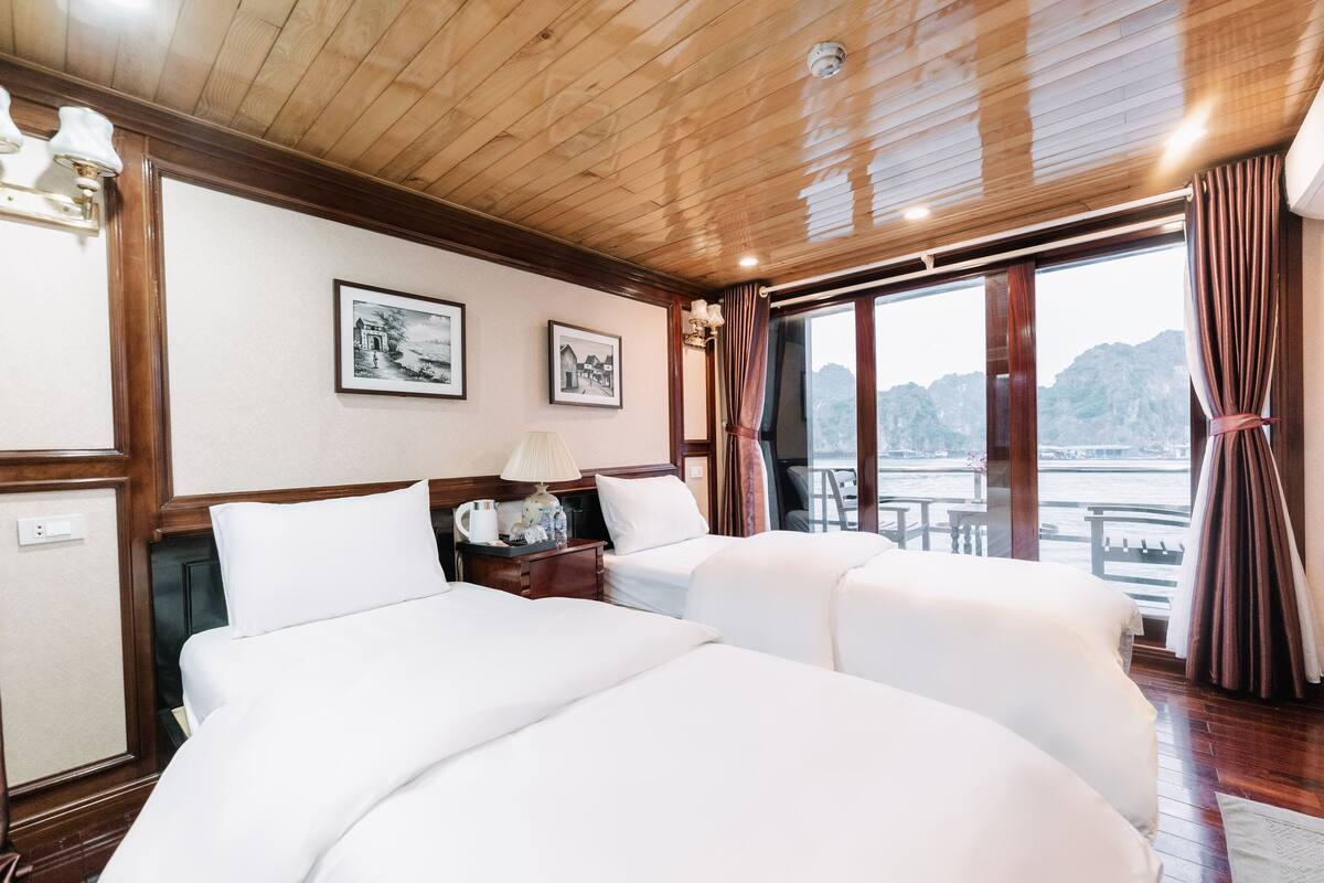 A hotel room with two beds, wooden ceiling, large windows with curtains, and framed pictures on the wall.