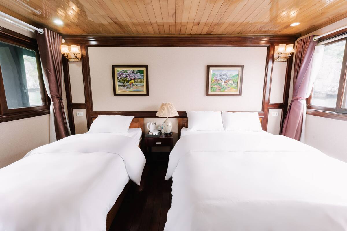 A hotel room with two beds, wooden ceiling, wall-mounted lamps, and two framed paintings.