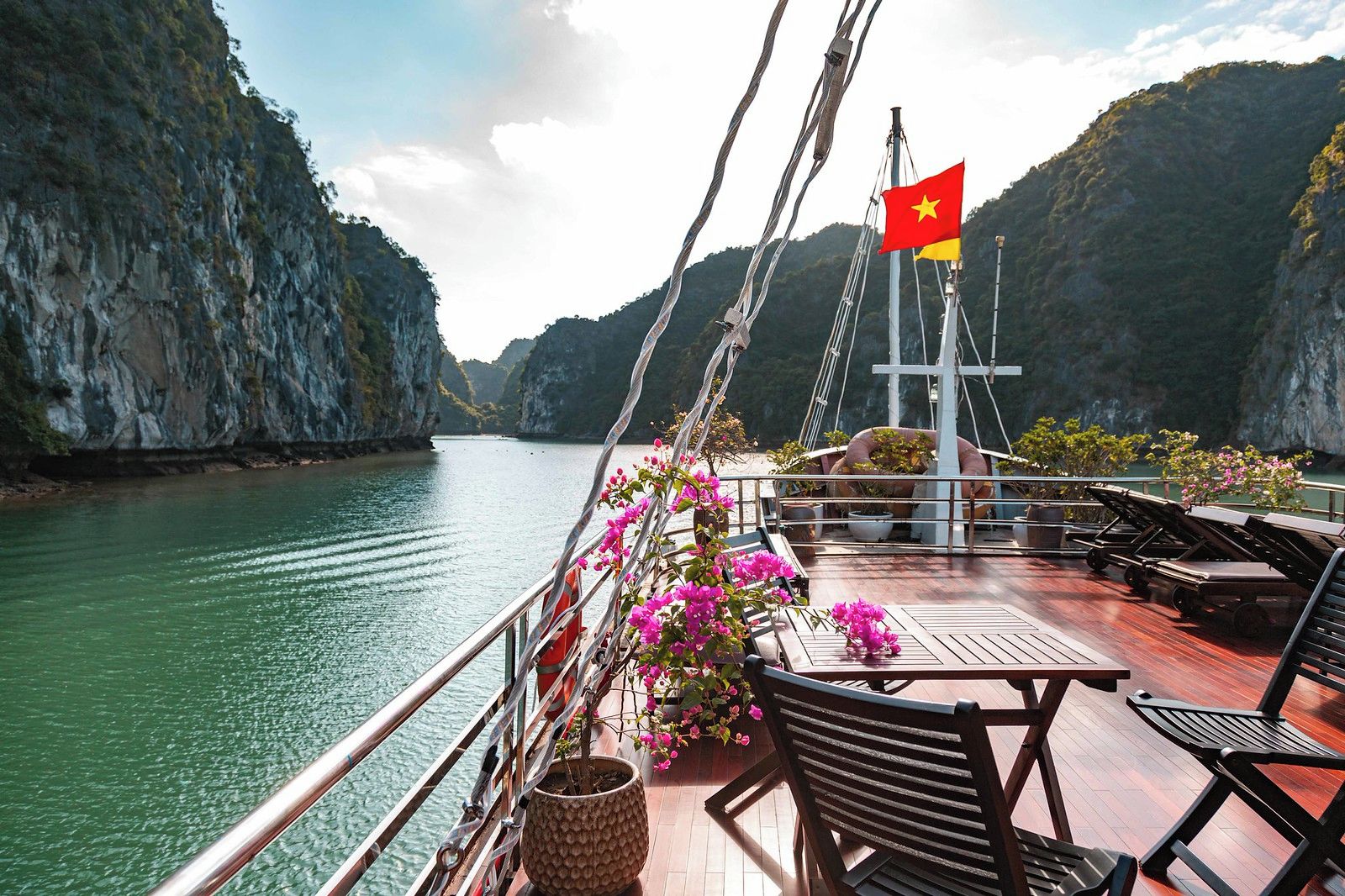 A boat deck with tables and chairs, a flag, and a view of a calm river surrounded by cliffs.