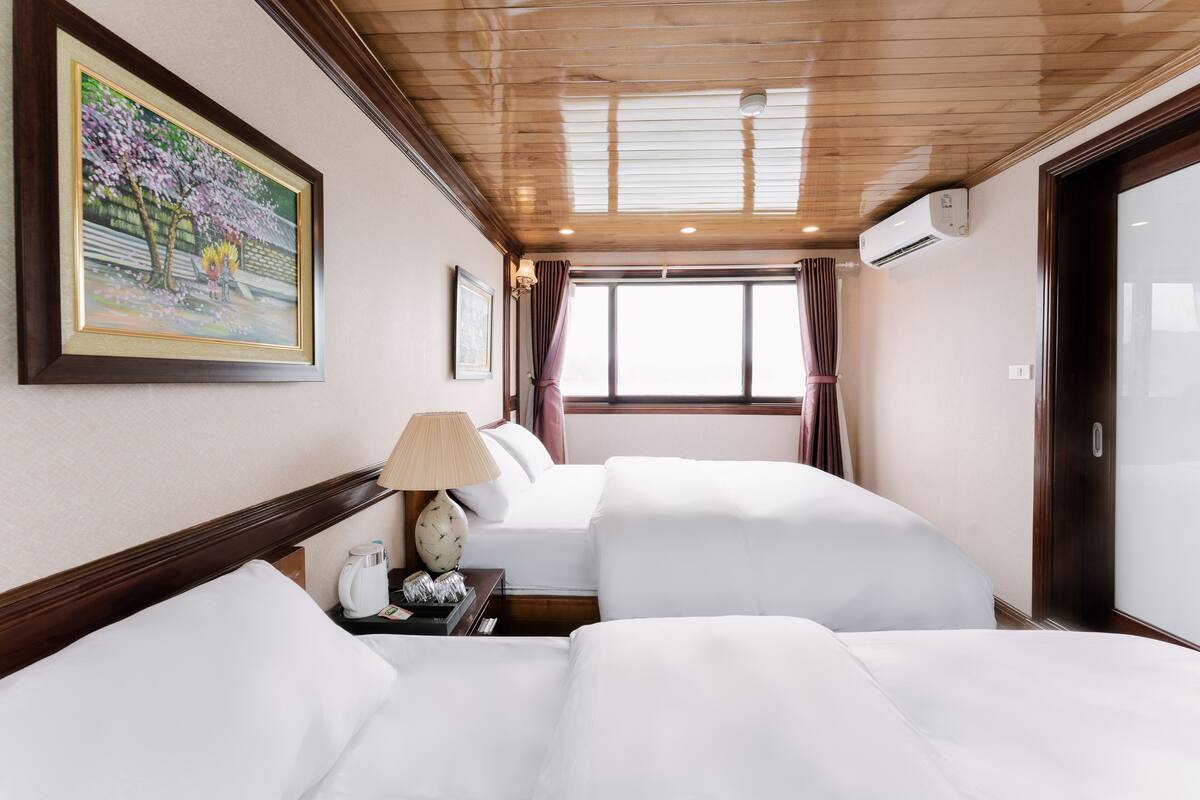 A hotel room with two beds, a painting on the wall, a window with curtains, and an air conditioning unit.