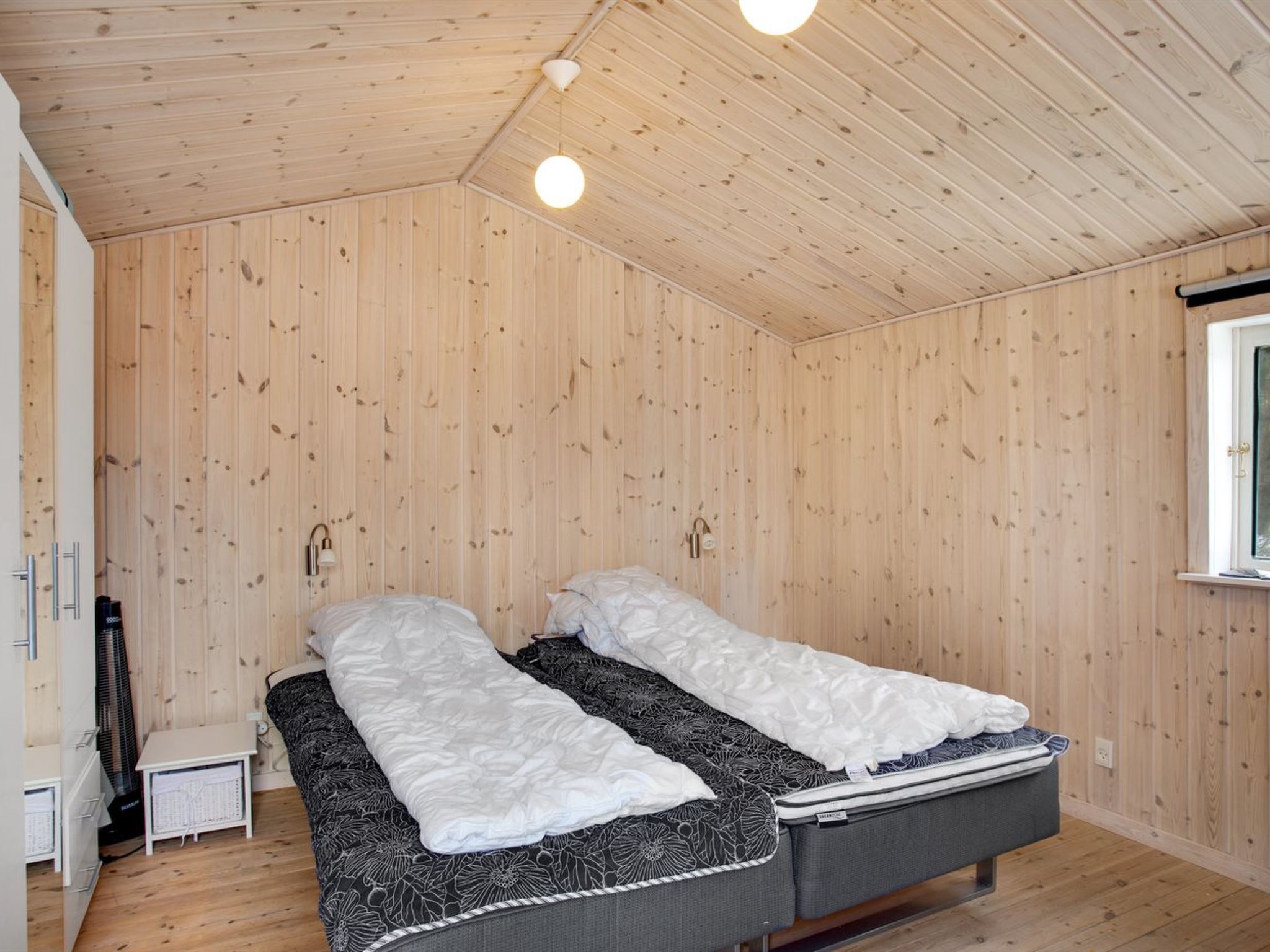 Ein Zimmer mit Holzwänden und -decke, zwei Einzelbetten mit weißer und gemusterter Bettwäsche, ein kleiner Nachttisch und ein Heizkörper.