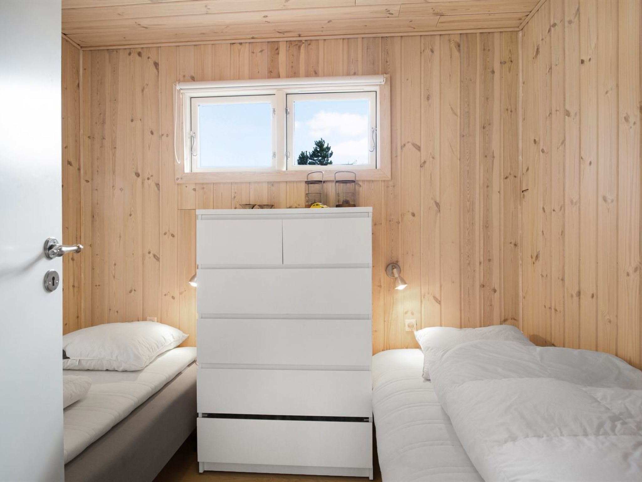 A room with wooden walls, a white dresser, two beds, and a window with a view of trees.