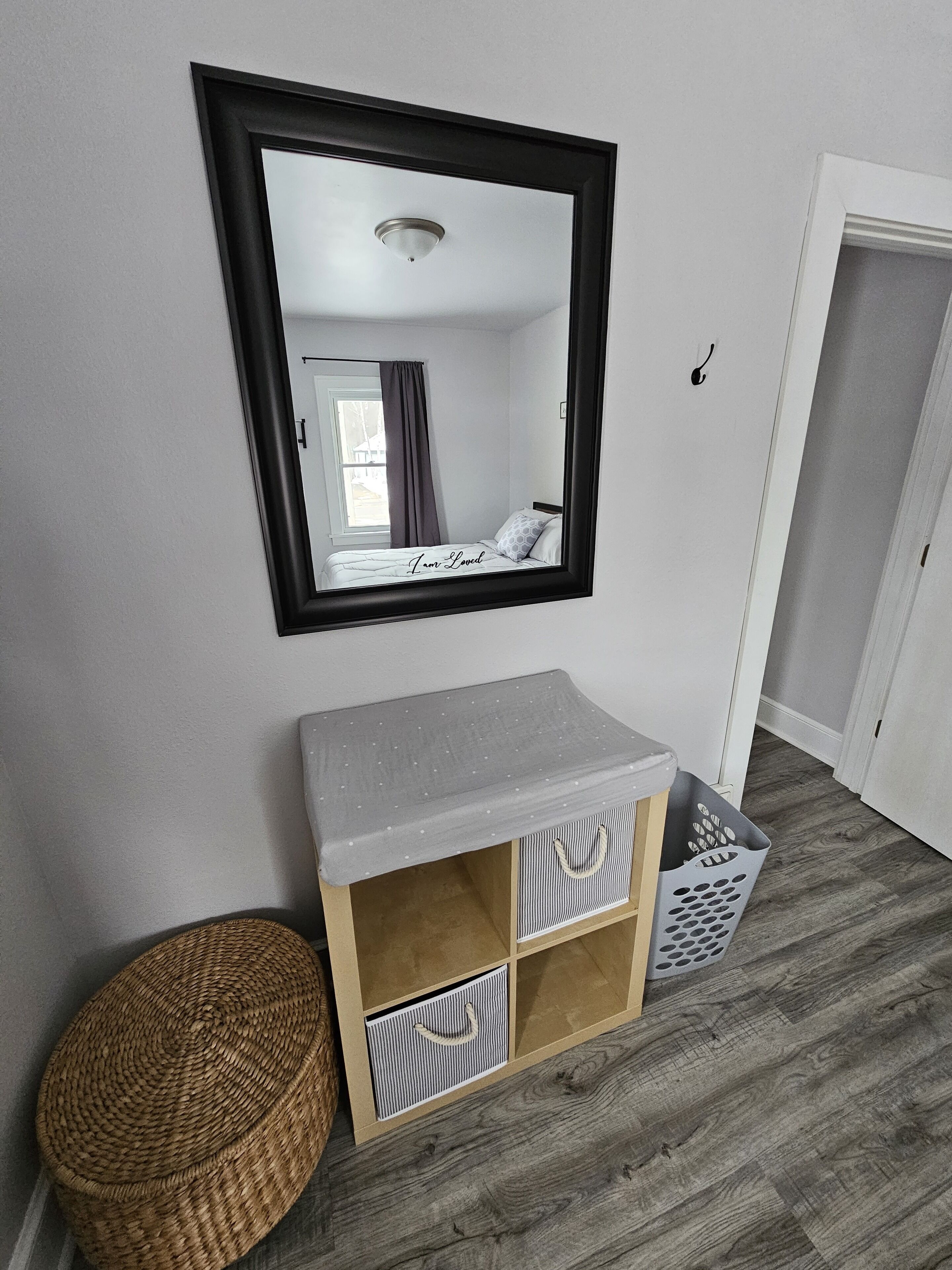 Una habitación con cama, espejo, un pequeño armario de madera, un taburete de mimbre y una puerta.