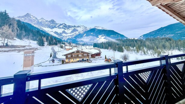 Ein Holzbalkon mit Blick auf eine verschneite Berglandschaft, im Hintergrund ein Chalet.