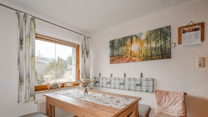 Ein Essbereich mit einem Holztisch, einer Bank und einem Stuhl. Ein Fenster mit Vorhängen ist sichtbar, und ein Waldgemälde hängt an der Wand.