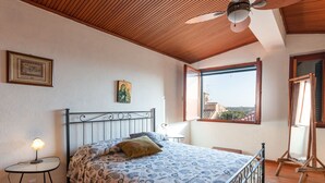 Una camera da letto con un letto, una ventola a soffitto, una finestra con vista, un tavolino e un quadro appeso al muro.