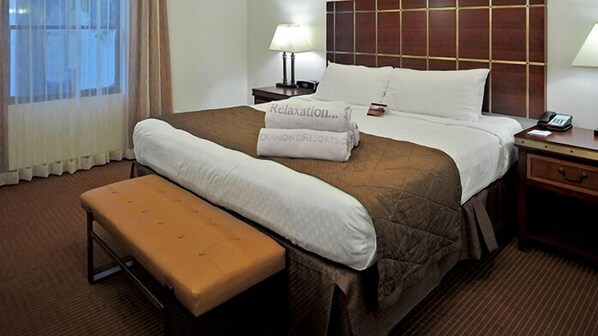 Habitación de hotel con cama, mesita de noche con lámpara, banco y ventana con cortinas.