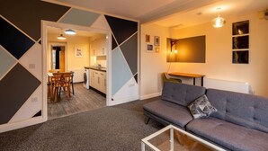 Een moderne woonkamer met een grijze bank en een glazen salontafel, een eethoek met een houten tafel en stoelen, en een keuken die zichtbaar is door een open deur.